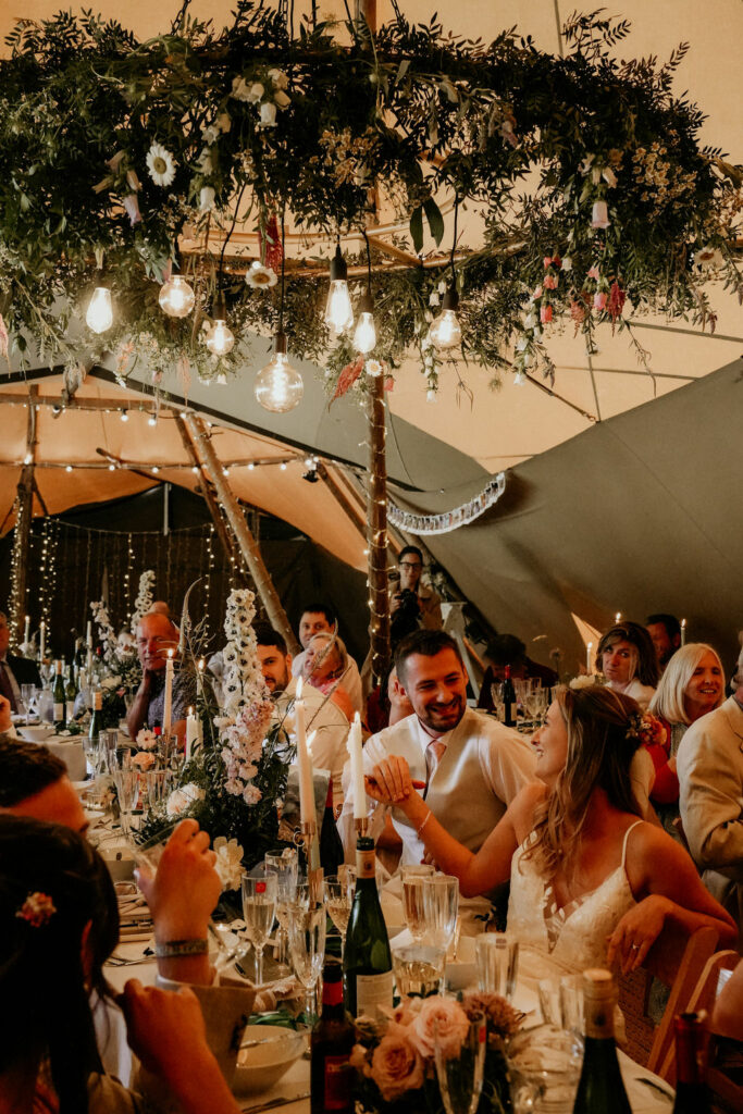 Tipi wedding guests with beautiful Edison chandelier covered in foliage. Created by wedding florist Winter and the Willows